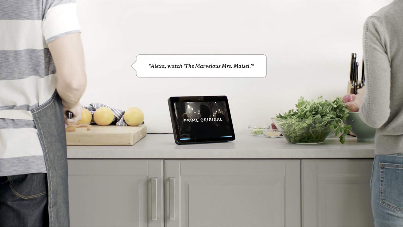  A man and a woman in kitchen. In the center of the picture is an Echo Show on a counter, with screen showing a movie frame and  texts reading as “Prime Original”  